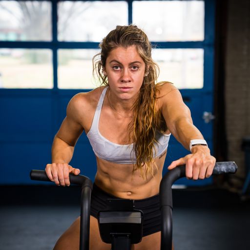 A woman on a fan bike, looking directly at the camera.  We can see her from the quadriceps and above.