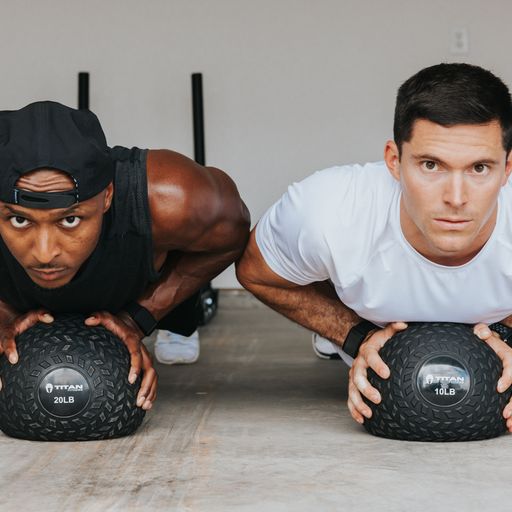 Two men doing pushups on slam balls, side by side, looking at the camera